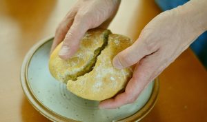 A pair of hands breaks a small bread roll over a handmade-style ceramic dish.