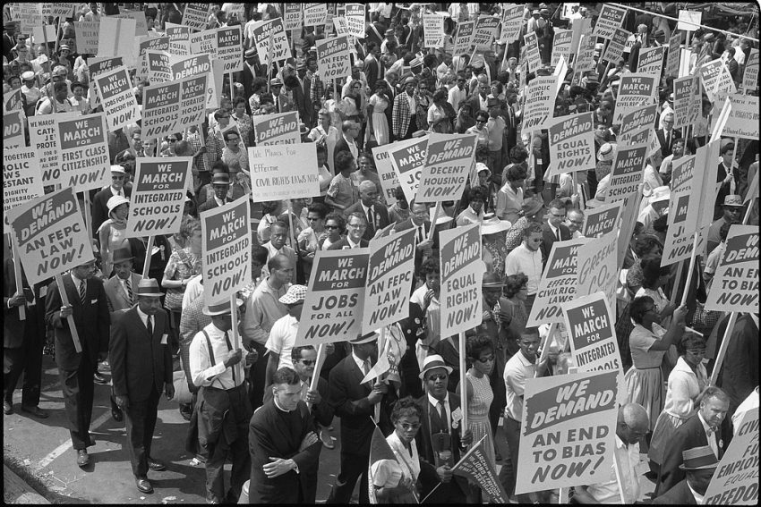 A black-and-white image of Demonstrators marching in the street holding signs during the March on Washington, 1963. The whole frame is filled with people walking forward; many are holding signs with sayings such as "We march for integrated schools NOW!" and "We march for jobs for all NOW!" and "We demand equal rights NOW!" and "We demand an end to bias NOW!"