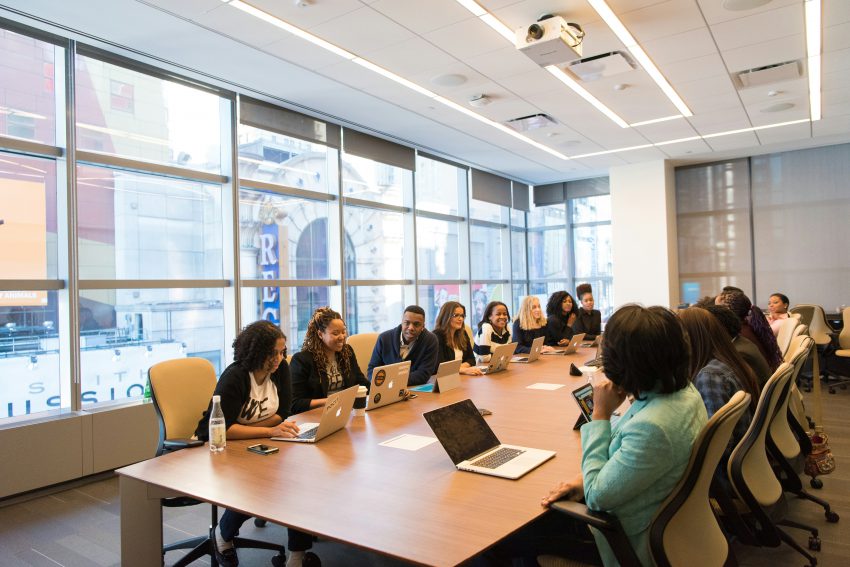 a happy looking group of people dressed for business gathers around a large table, prepared for a meeting
